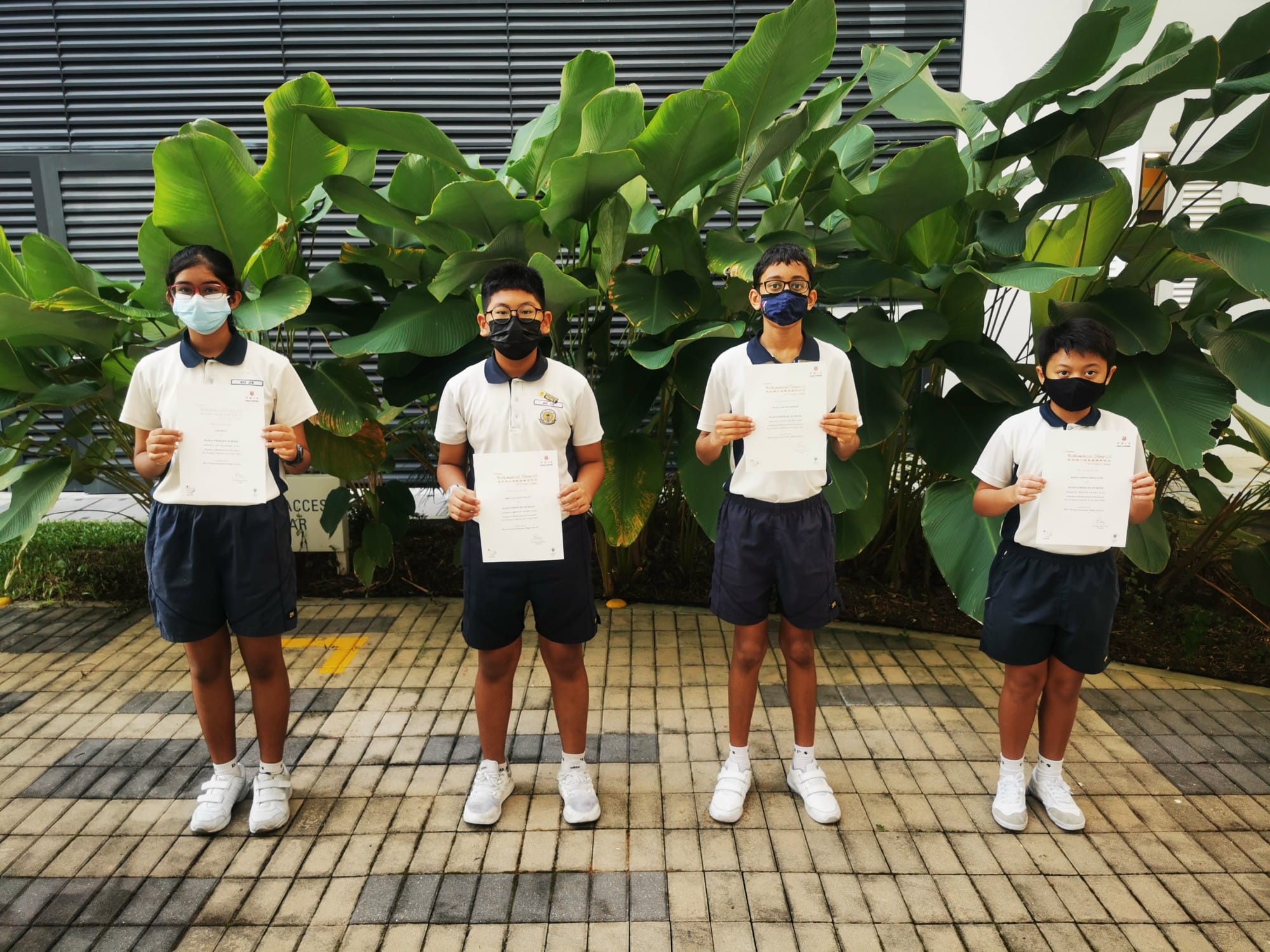 Four masked students in white and navy uniforms stand outdoors holding certificates in front of large green leaves.